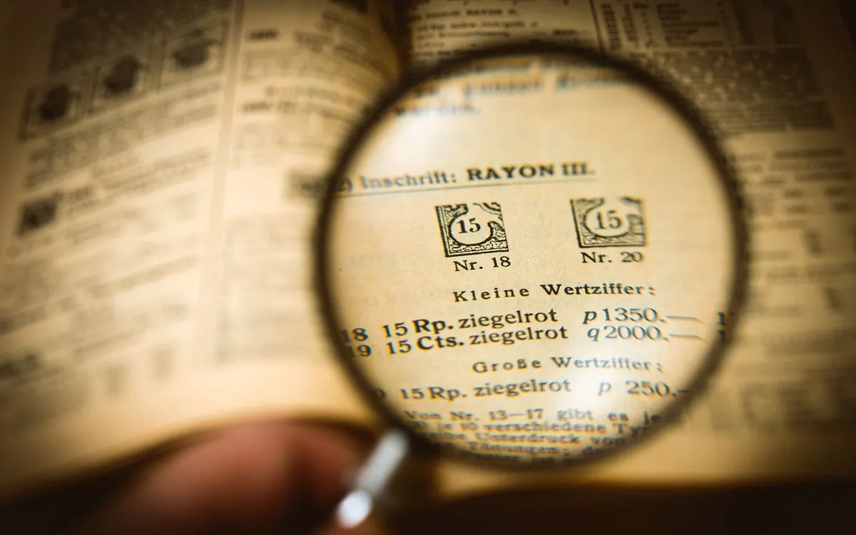 A magnifying glass held over a printed stamp catalogue, enlarging a row of Rayon III entries priced in Rappen and Centimes.