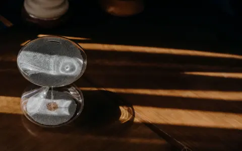 A small glass sand timer resting on a dark wooden table in low warm light.