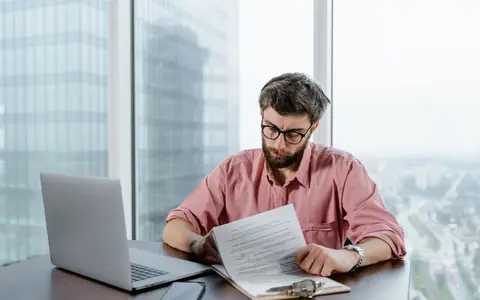 A man in glasses reading printed pages at a desk beside a floor to ceiling office window, with an open laptop beside him.