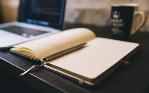 An open blank notebook with a pen across it, a laptop and a mug out of focus behind.