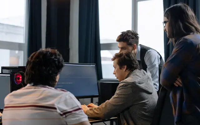 Four people gathered around a desktop computer in a modern office, watching the screen together.