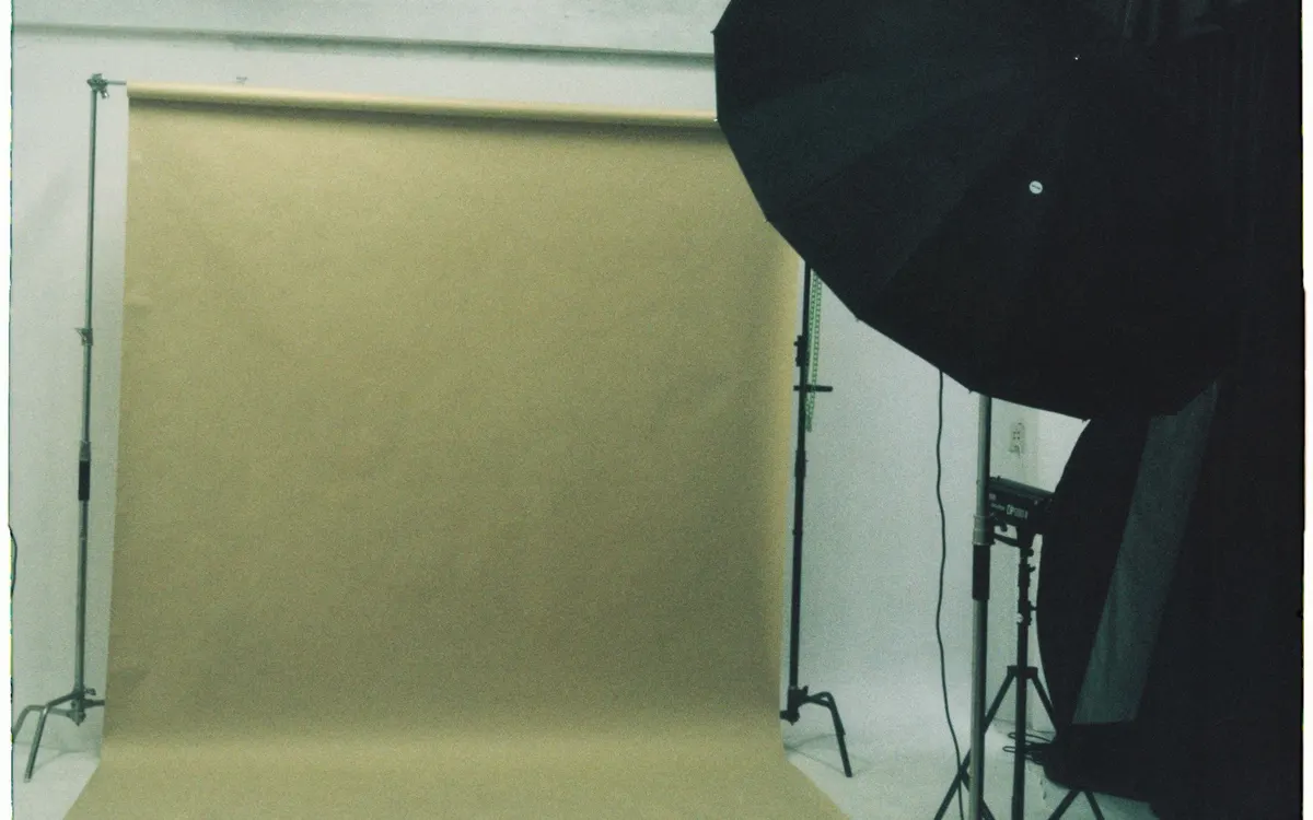 A photography studio with a beige paper backdrop mounted on stands and black lighting equipment with a softbox on a tripod.