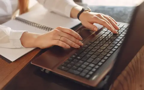 A person types on a black laptop keyboard at a wooden desk beside a spiral notebook in warm light.