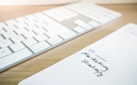 A white wireless keyboard rests on a light wooden desk beside an open notebook with handwritten notes in natural light.