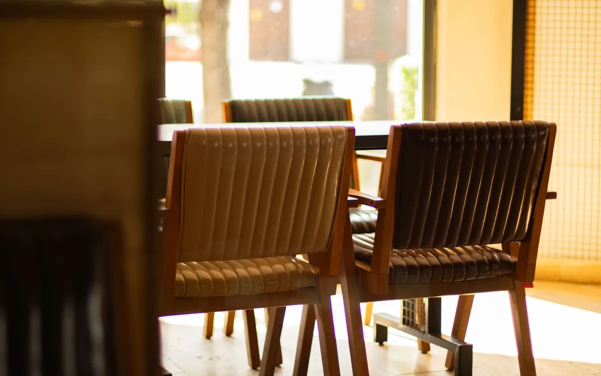 Chairs with leather seats and wooden frames arranged around a table in a bright interior with large windows and warm natural light.