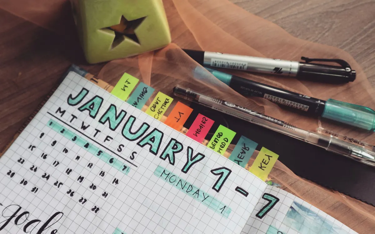 A handwritten planner with colorful sticky notes, gel pens and a coffee mug on a wooden desk.